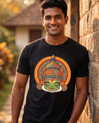 A man wearing a black t-shirt with a colorful traditional Indian theatrical mask graphic, standing against a rustic stone wall.