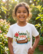 A young girl smiling in a sunny garden, wearing a white t-shirt with a colorful graphic that reads 'ONAM VIBES' above a traditional boat illustration.