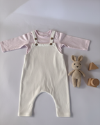 Baby outfit with pink shirt and beige overalls on a light gray background, accompanied by a plush toy and wooden blocks.