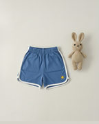Blue shorts with white trim and a small logo, paired with a knitted bunny toy on a light gray background.