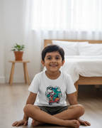 Child sitting on the floor in a bedroom wearing a white t-shirt with a graphic design.