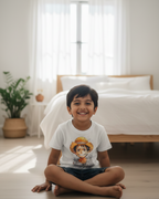 Child sitting on the floor wearing a white t-shirt with a graphic design, in a bright room with a bed and plants.