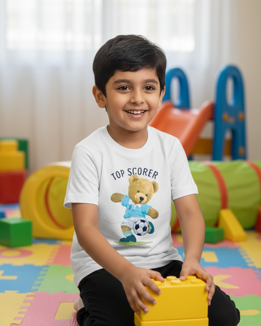 Child wearing a 'Top Scorer' t-shirt with teddy bear and soccer ball design, sitting on colorful foam mats.
