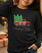 Child wearing a black long-sleeve shirt with a Christmas-themed design, standing in front of a decorated Christmas tree and presents.