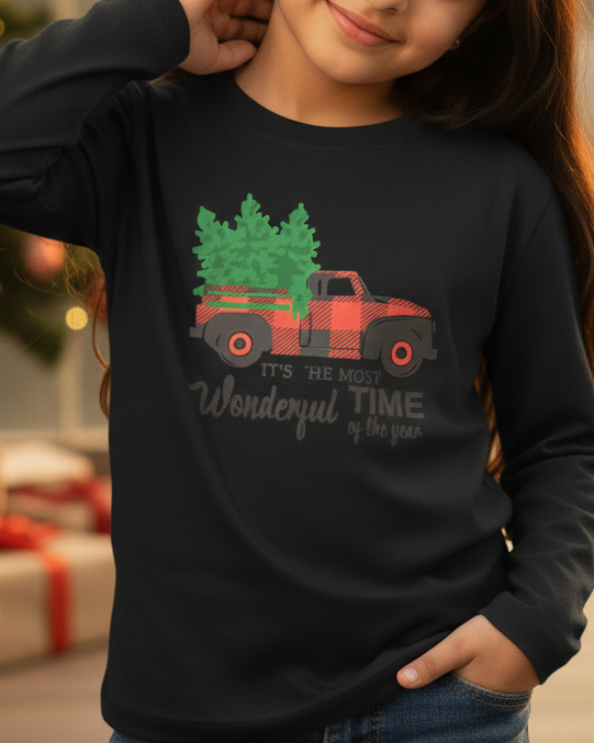 Child wearing a black long-sleeve shirt with a Christmas-themed design, standing in front of a decorated Christmas tree and presents.