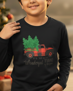 Child wearing a black long-sleeve shirt with a Christmas tree and truck design, standing in front of a decorated Christmas tree.