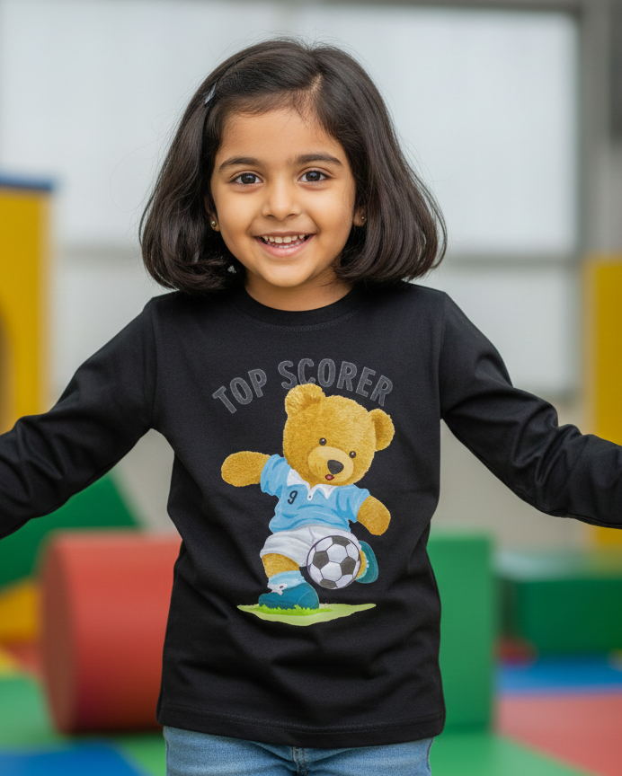 Child wearing a black long-sleeve shirt with a teddy bear graphic and 'Top Scorer' text in a colorful indoor setting.