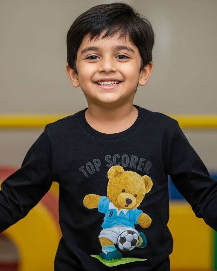 Child wearing a black long-sleeve shirt with a teddy bear graphic and 'Top Scorer' text, standing in an indoor play area.
