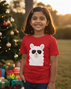 Child wearing a red shirt with a llama graphic in front of a Christmas tree and presents.