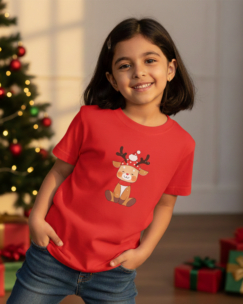 Child wearing a red t-shirt with a reindeer design in a festive setting with Christmas tree and presents.