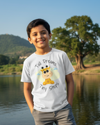 Child wearing a t-shirt with a cartoon character and text, standing by a lake with trees and mountains in the background.