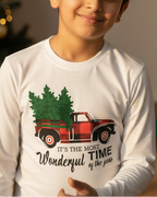Child wearing a white long-sleeve shirt with a Christmas design, standing in front of a decorated Christmas tree and presents.