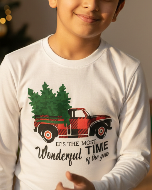 Child wearing a white long-sleeve shirt with a Christmas design, standing in front of a decorated Christmas tree and presents.