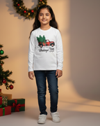 Child wearing a white long-sleeve shirt with a Christmas design, standing next to a decorated Christmas tree and presents.