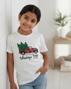 Child wearing a white t-shirt with a Christmas truck design in a room with a white wall and wooden shelf.