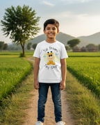 Child wearing a white t-shirt with a cartoon character and text, standing on a dirt path in a green field with trees and mountains in the background.