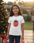 Child wearing a white t-shirt with a red and black ladybug design, standing in front of a decorated Christmas tree and presents.