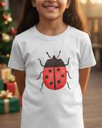 Child wearing a white t-shirt with a red ladybug design, standing in front of a decorated Christmas tree.