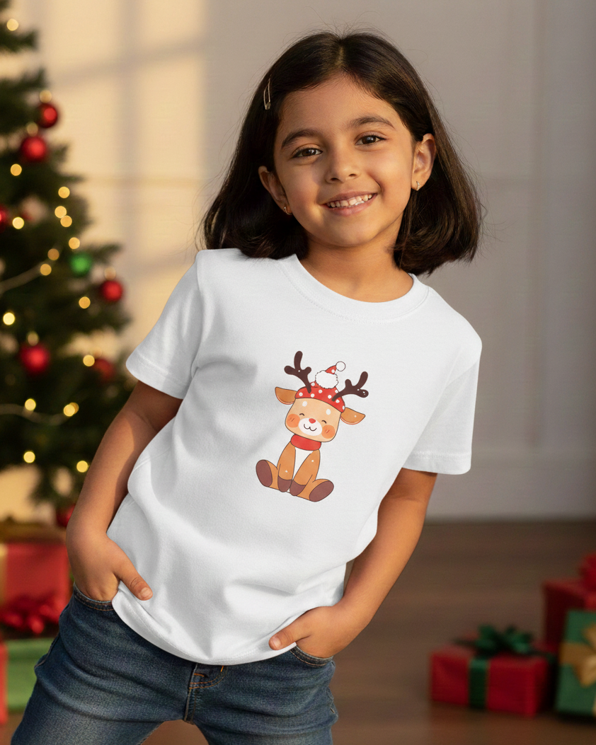 Child wearing a white t-shirt with a reindeer design in a festive setting with Christmas tree and presents.