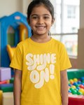 Child wearing a yellow shirt with 'SHINE ON!' text in a colorful indoor setting