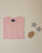 Folded pink t-shirt with a brand label on a light beige background, accompanied by wooden rings and star-shaped objects.