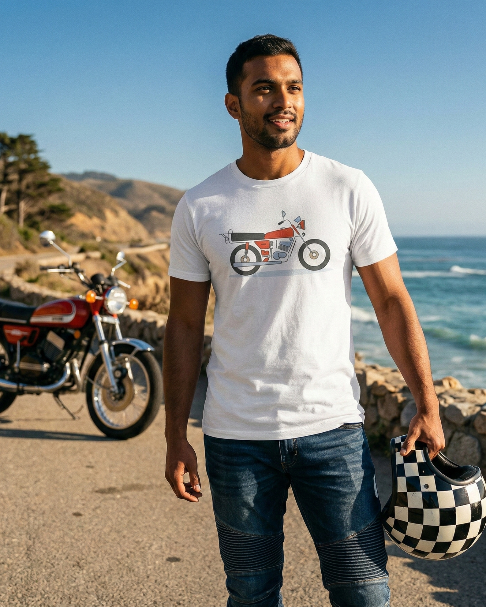 Man standing on a road by the ocean with a motorcycle and helmet, wearing a t-shirt with a motorcycle graphic.