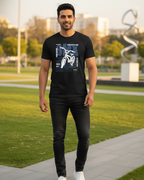 Man wearing a black t-shirt with a graphic design, standing outdoors on a path with greenery and a sculpture in the background.