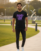 Man wearing a black t-shirt with purple text, standing on a path with greenery in the background