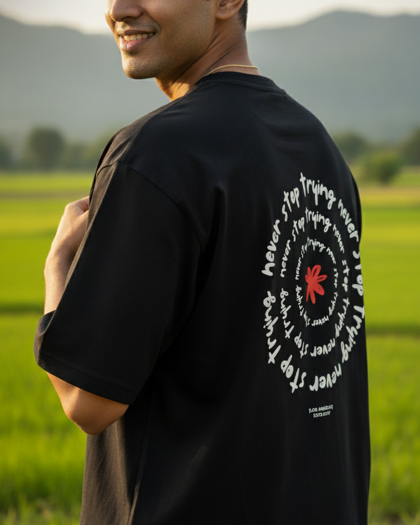 Man wearing a black t-shirt with text and a red circle design, standing in a green field with mountains in the background.