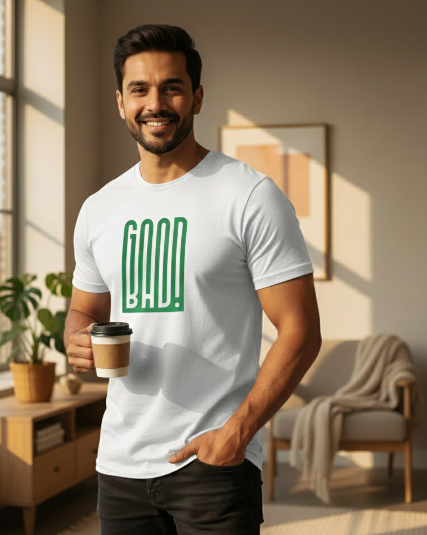 Man wearing a white t-shirt with 'GOOD BAD' in green, holding a coffee cup in a room.