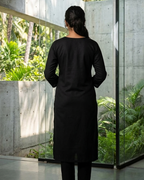 Person wearing a black outfit standing in a modern architectural setting with greenery.
