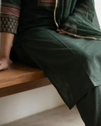 Person wearing a green traditional outfit with patterned cuffs sitting on a wooden bench.