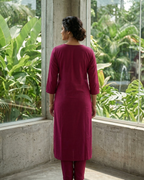 Person wearing a maroon outfit standing in front of a glass wall with greenery outside.