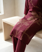 Person wearing a maroon outfit with gold embroidery sitting on a light wooden bench.