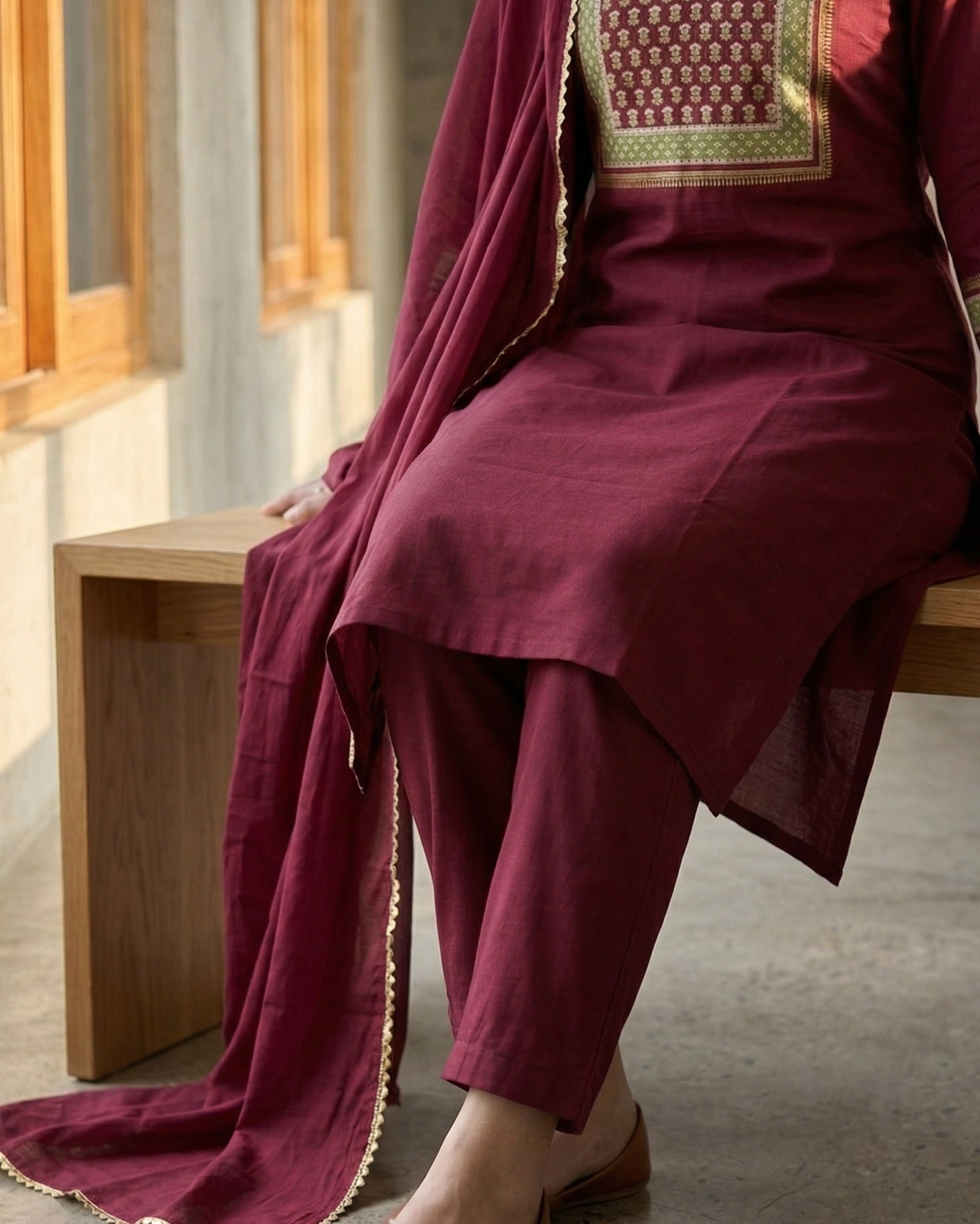 Person wearing a maroon traditional outfit with gold embroidery sitting on a wooden bench.