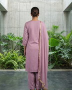 Person wearing a mauve traditional outfit with a gold border in an outdoor setting with plants and concrete walls.