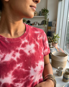 Person wearing a pink and white tie-dye shirt in a room with plants and a shelf.