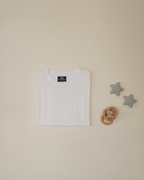 White baby t-shirt with a brand label on a beige background, accompanied by wooden rings and star-shaped toys.