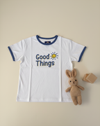 White t-shirt with 'Good Things' text and sun graphic, accompanied by a plush rabbit and wooden block on a beige background.