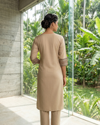 Woman in a beige outfit standing in front of a glass wall with a view of greenery.
