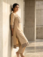 Woman in a beige traditional outfit standing against a light-colored wall.