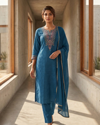 Woman in a blue traditional outfit standing in a modern indoor setting with large windows.