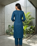 Woman in a blue traditional outfit standing in a modern indoor setting with plants.