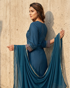 Woman in a blue traditional outfit with a matching dupatta against a textured wall.
