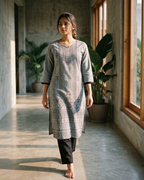 Woman in a gray patterned kurta standing in a sunlit room with large windows and plants.