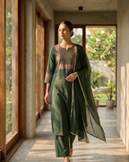 Woman in a green traditional outfit standing in a sunlit room with large windows.