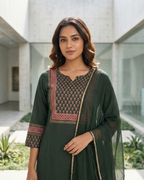 Woman in a green traditional outfit standing in an outdoor setting with modern architecture.