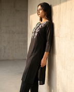 Woman wearing a black outfit with floral embroidery leaning against a wall.