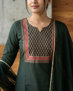 Woman wearing a dark green traditional outfit with red and gold patterns against a wooden wall.
