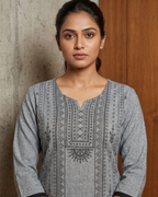 Woman wearing a gray patterned shirt against a wooden and stone wall.